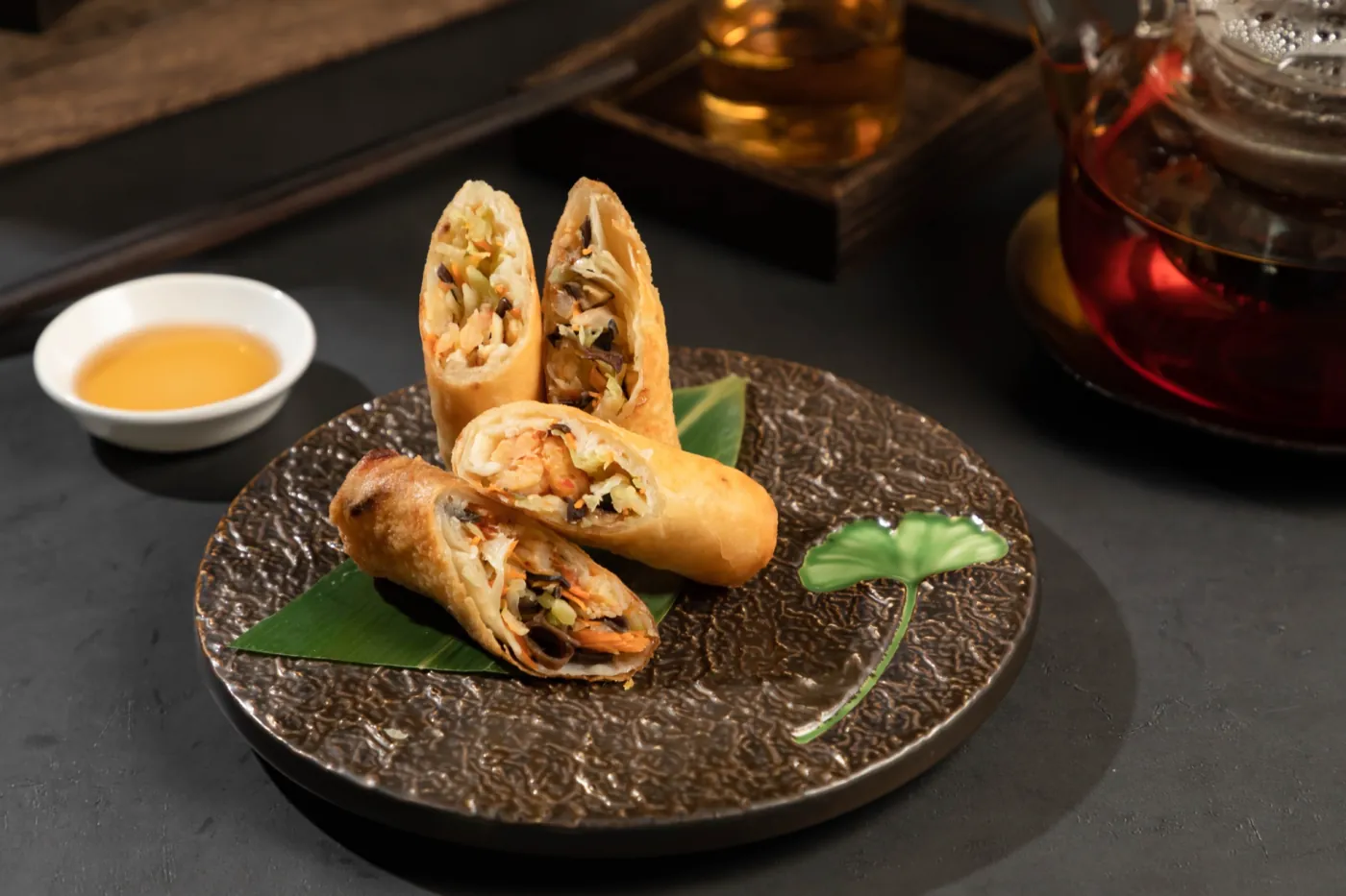 Golden spring rolls, halved to show their filling, on a dark hammered plate with dipping sauce
